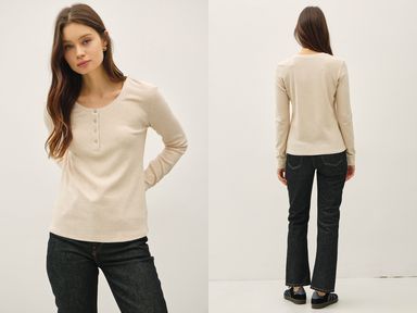 Woman wearing a beige long-sleeve top and dark jeans, standing against a white background.