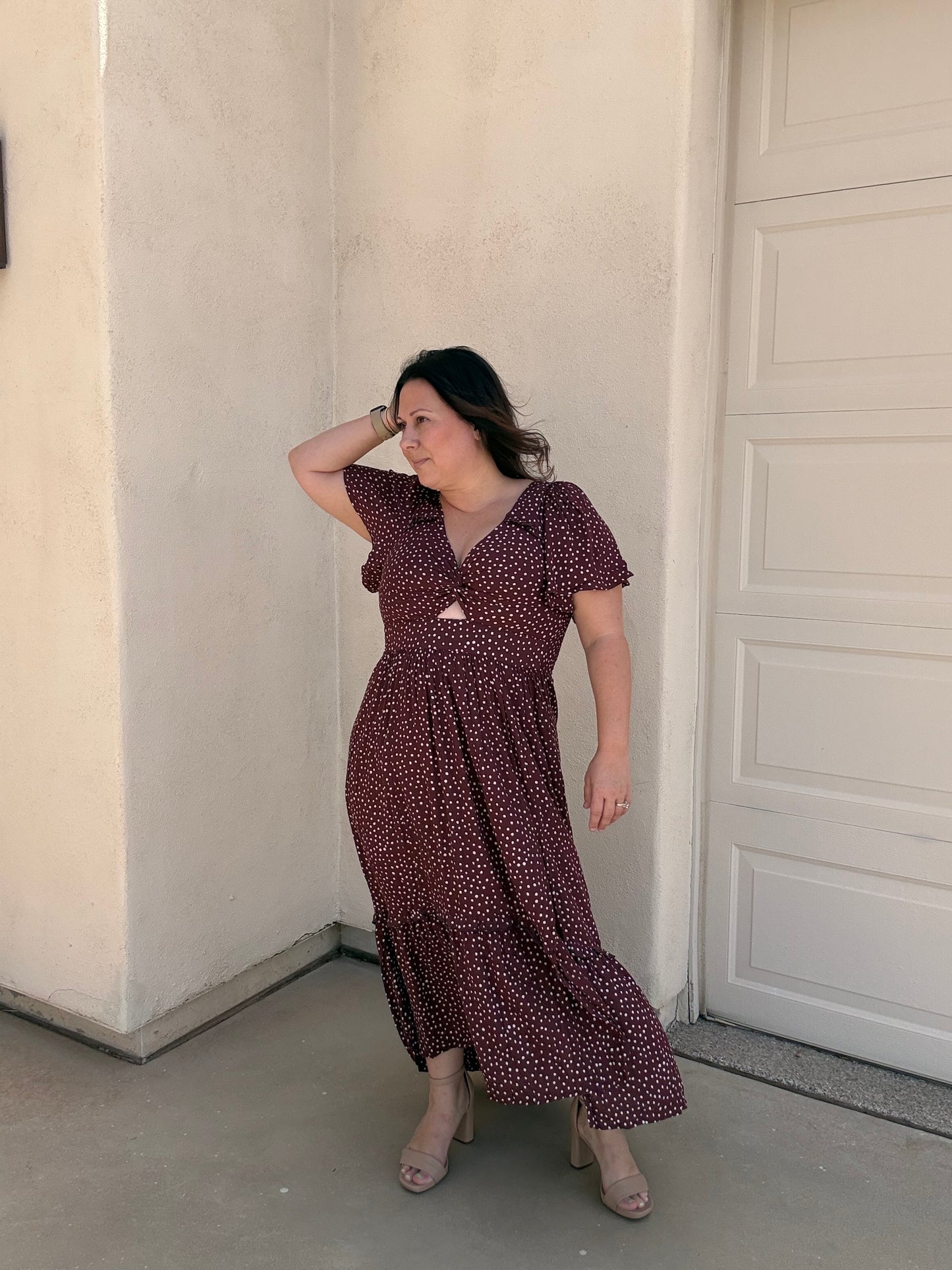 A woman wearing a dark burgundy maxi dress with a small white floral pattern, standing in front of a beige stucco wall and a white garage door.