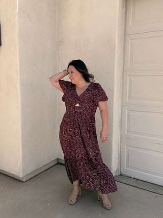 A woman wearing a dark burgundy maxi dress with a small white floral pattern, standing in front of a beige stucco wall and a white garage door.