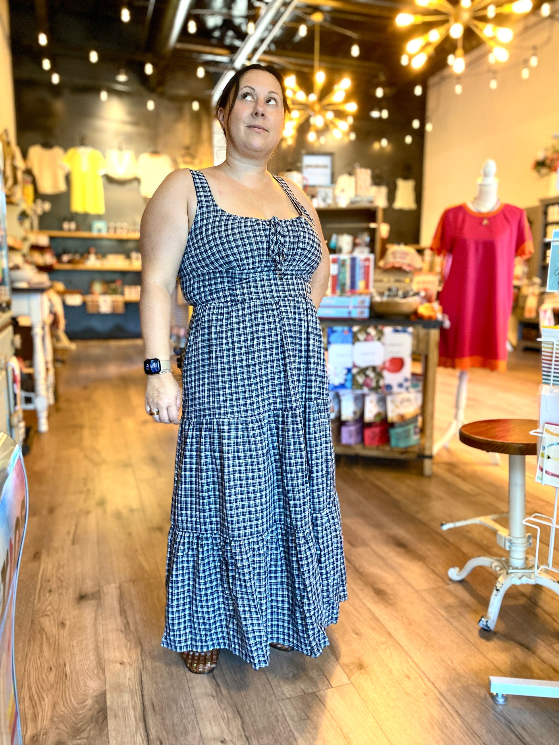 A woman wearing a blue and white gingham tiered maxi dress stands in a boutique store with wood floors and warm hanging lights.