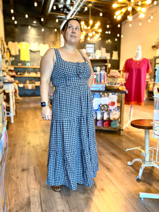 A woman wearing a blue and white gingham tiered maxi dress stands in a boutique store with wood floors and warm hanging lights.
