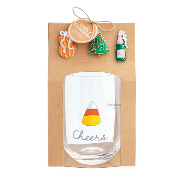 A clear glass tumbler with a candy corn graphic and the word 'Cheers' printed on it, presented inside a brown kraft gift box. The box is decorated with three small enamel pins (a pumpkin, a Christmas tree, and a champagne bottle) and a hanging tag that reads 'Simply Celebrate'.