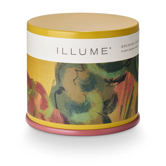 A cylindrical candle tin by ILLUME with a mustard yellow lid, a white band with the brand name, and an abstract painted label in green, pink, and yellow. The tin sits on a pink base against a clean white background.