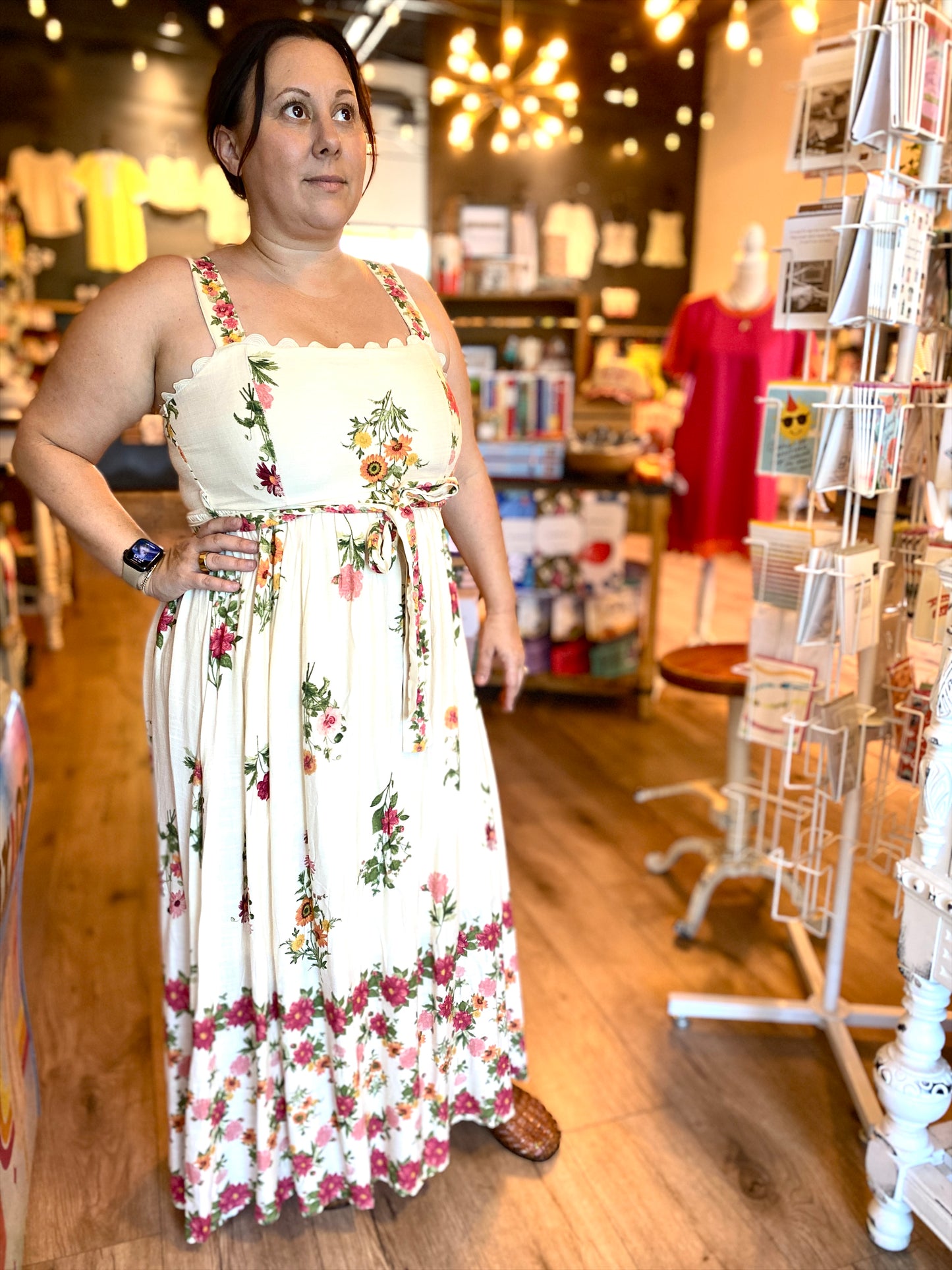A woman wearing a white sleeveless maxi dress with a colorful floral print and scalloped neckline, standing inside a retail store with wooden floors and display racks.