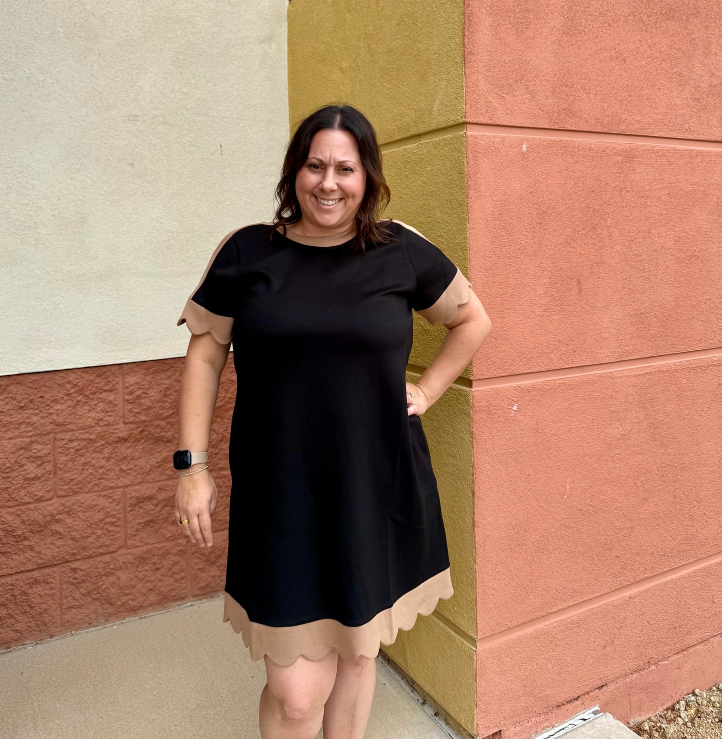 A woman wearing a black short-sleeve dress with beige scalloped trim on the sleeves and hem, standing outdoors against a textured wall corner.