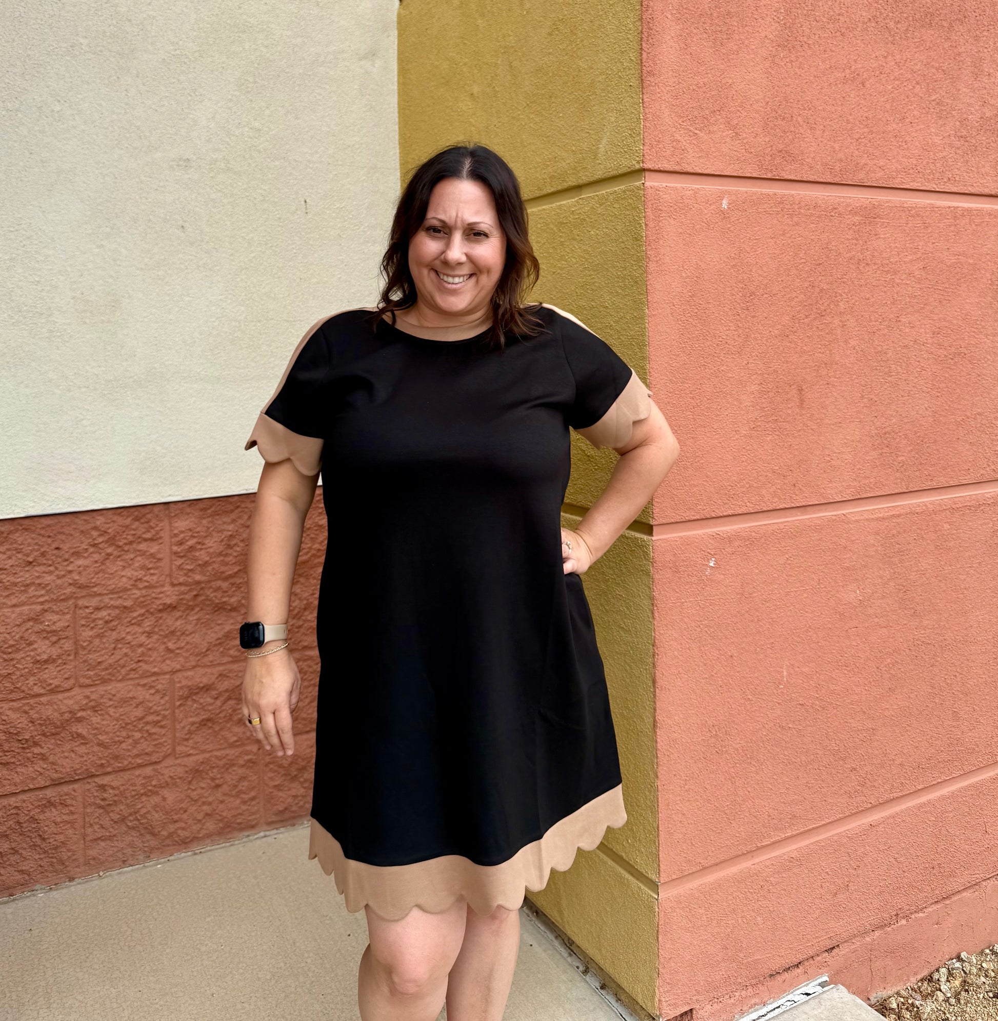 A woman wearing a black short-sleeve dress with beige scalloped trim on the sleeves and hem, standing outdoors against a textured wall corner.