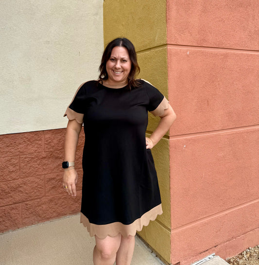 A woman wearing a black short-sleeve dress with beige scalloped trim on the sleeves and hem, standing outdoors against a textured wall corner.
