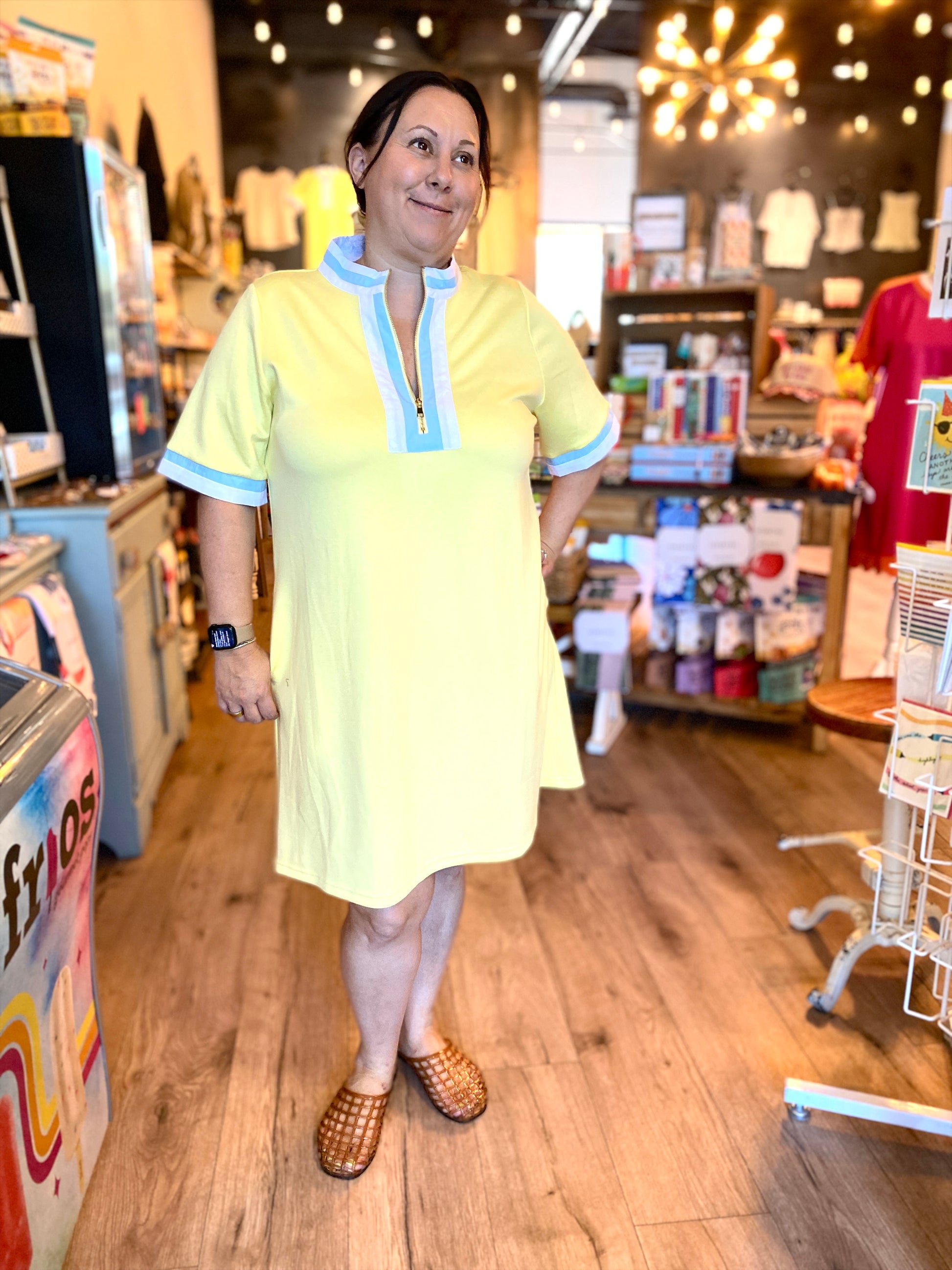 A woman wearing a pale yellow short-sleeve tunic dress with light blue trim stands inside a retail store with wood floors and shelves in the background.