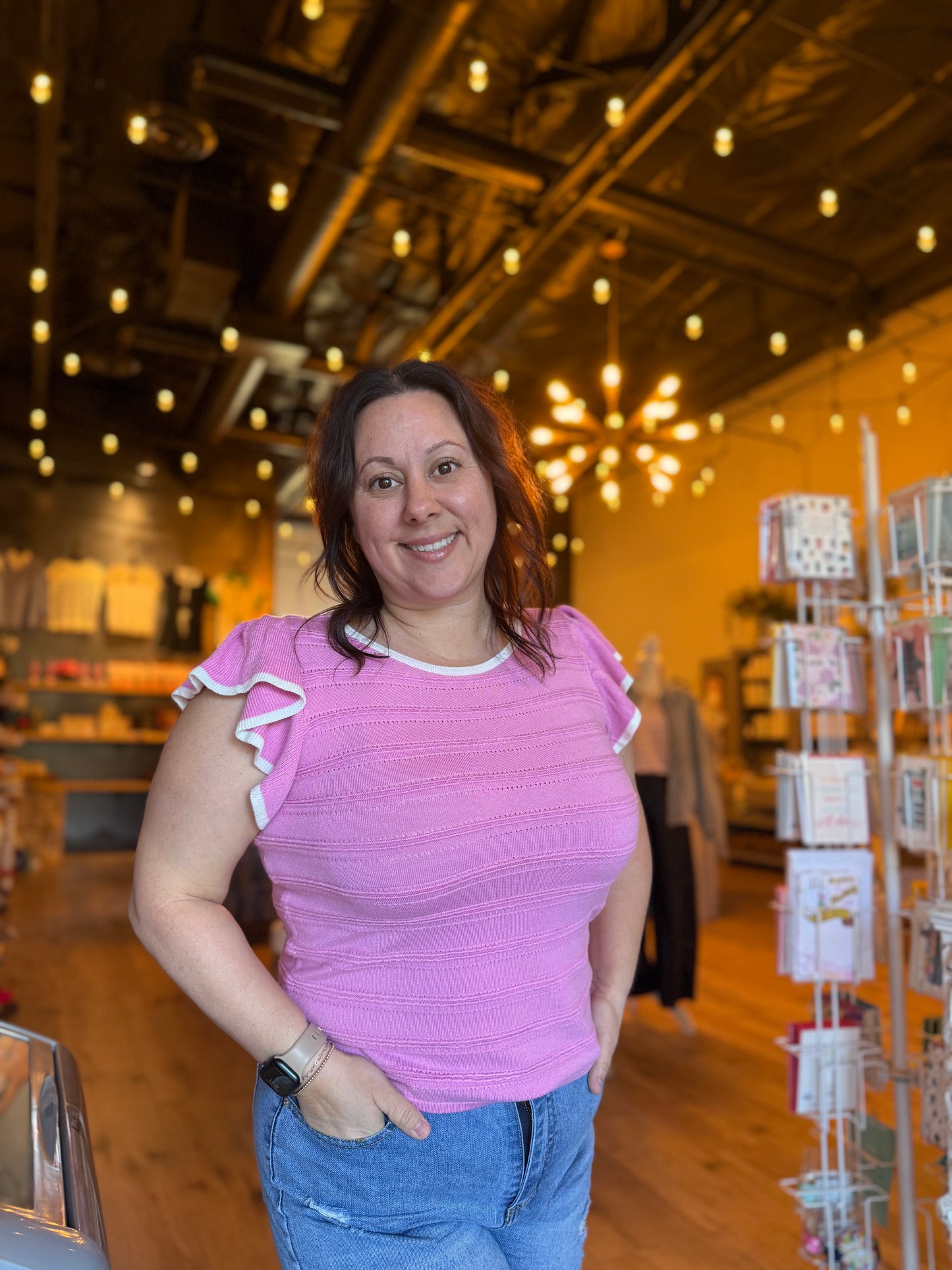 Woman in a pink shirt standing in a store with wooden ceiling and card display.