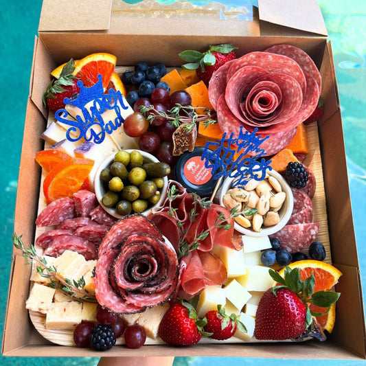 Gourmet charcuterie board with meats, cheeses, fruits, and vegetables in a cardboard box with 'Super Dad' sign.