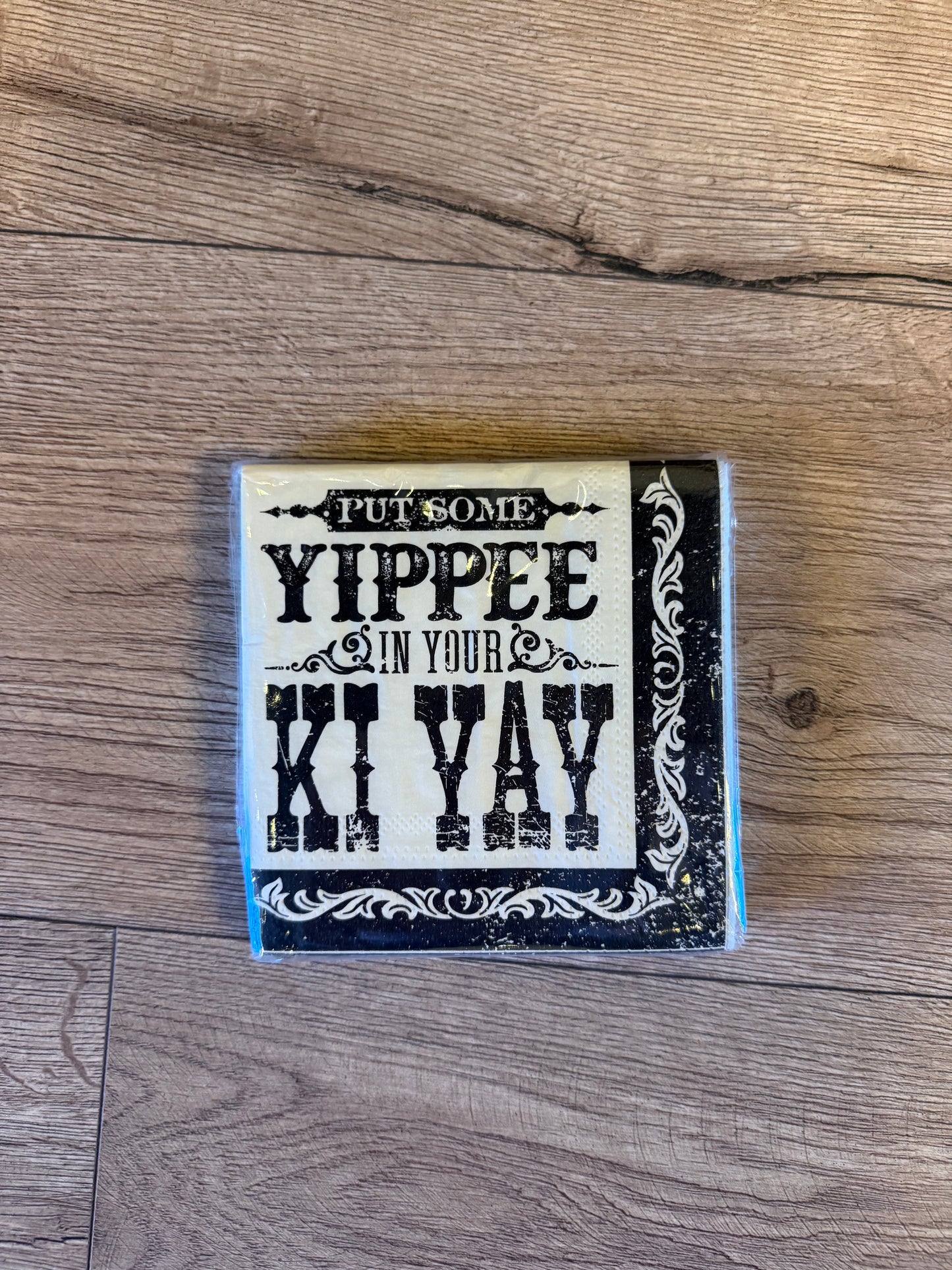 A square decorative sign with a distressed black and cream design featuring the text 'PUT SOME YIPPEE IN YOUR KI YAY', placed on a light brown wood floor.