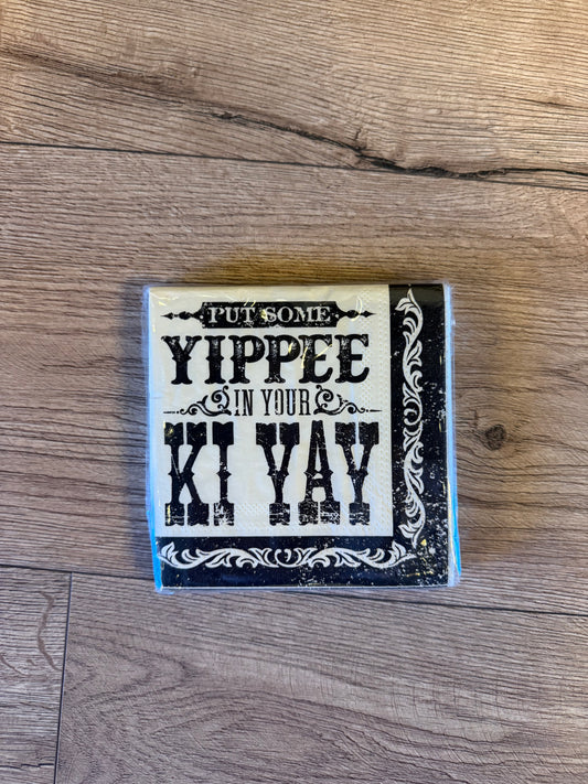 A square decorative sign with a distressed black and cream design featuring the text 'PUT SOME YIPPEE IN YOUR KI YAY', placed on a light brown wood floor.