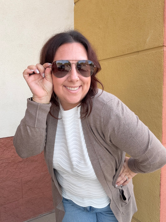 Woman wearing sunglasses and a taupe sweater against a multicolored wall.