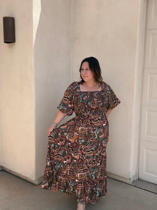 A woman wearing a bohemian-style maxi dress with a complex paisley and floral pattern in brown, rust, orange, and sage green, standing against a beige stucco wall.