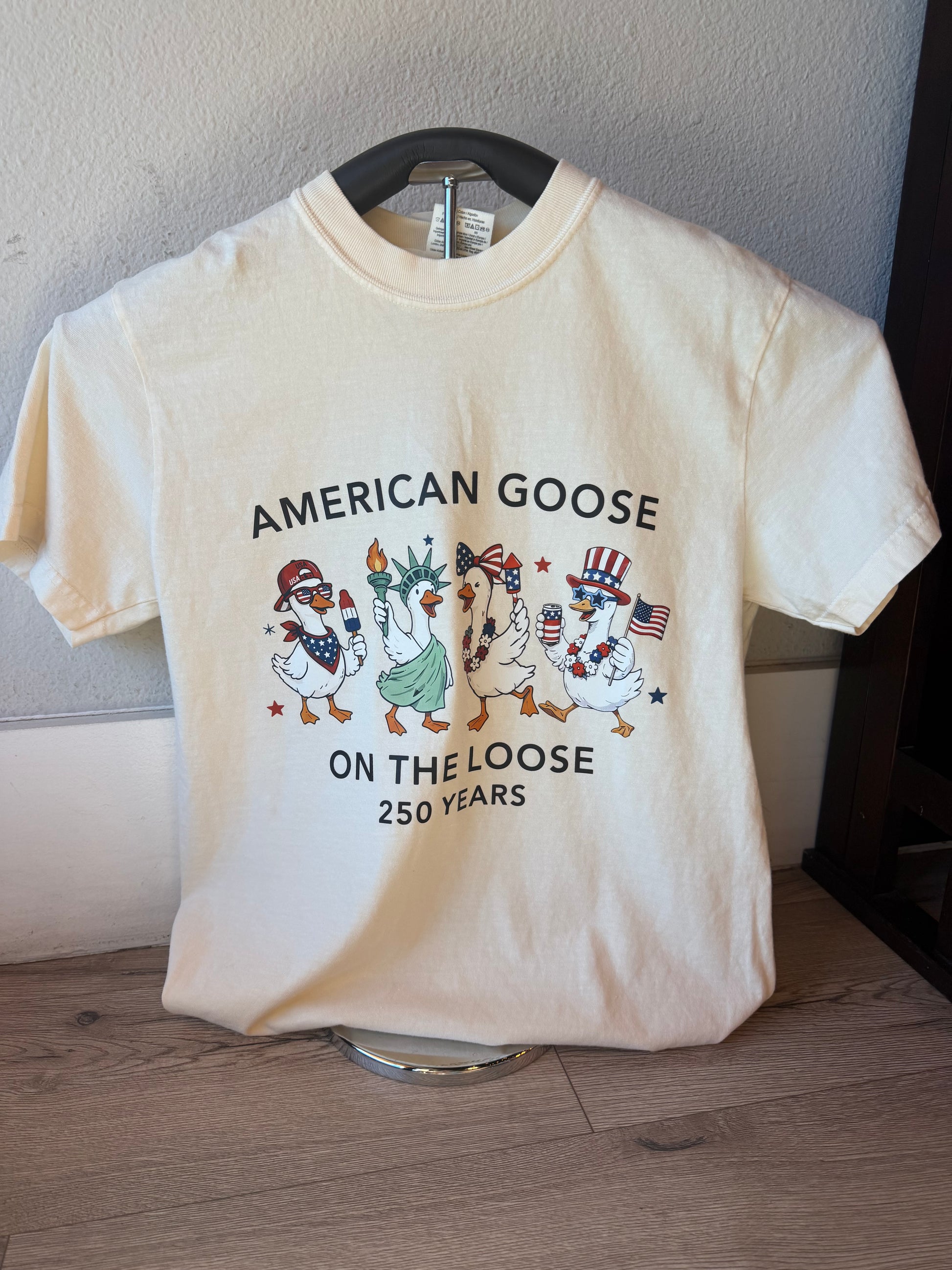 A cream-colored short-sleeve t-shirt on a hanger, featuring a graphic of four cartoon American geese with patriotic hats and the text 'AMERICAN GOOSE ON THE LOOSE 250 YEARS'.