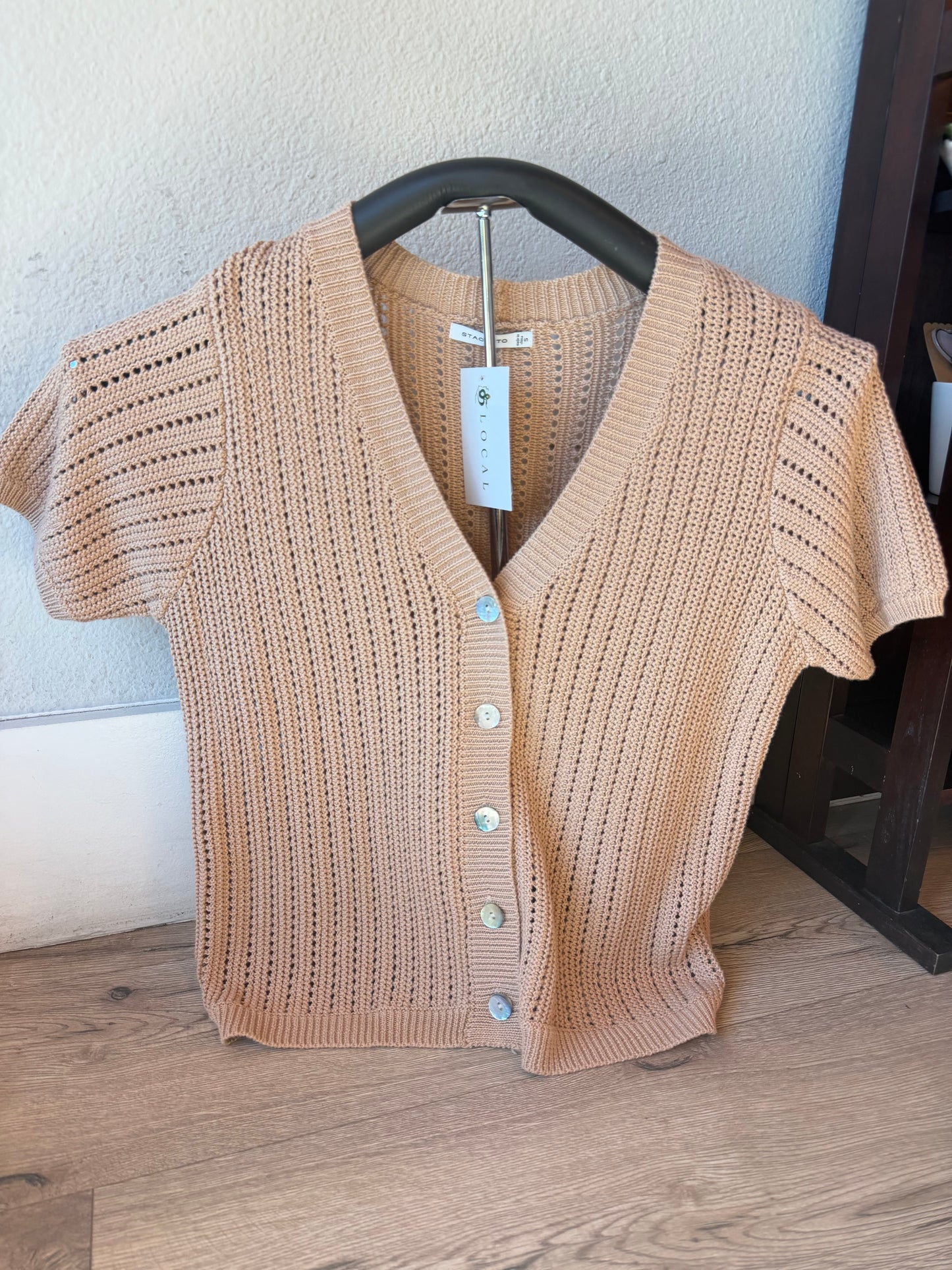 A beige short-sleeve knit cardigan sweater with a V-neck and decorative buttons, hanging on a black hanger against a light wall and wood floor.