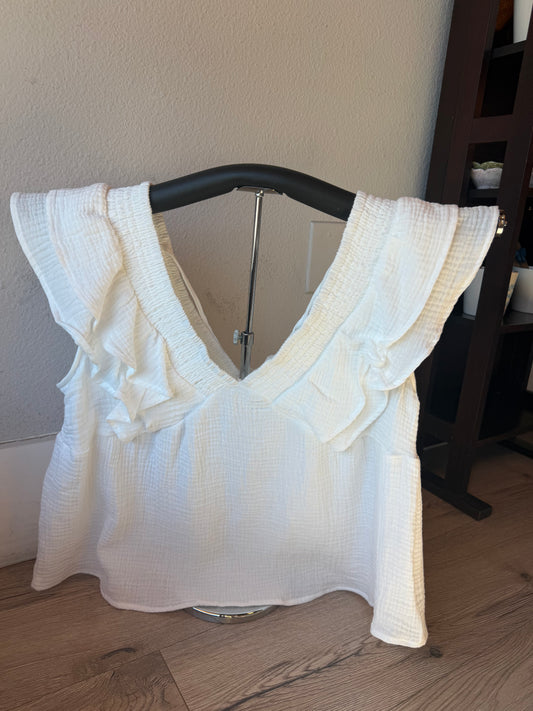 A white textured cotton top with ruffle details on the shoulders and a deep V-neckline, displayed on a black hanger on a wooden floor against a beige wall.