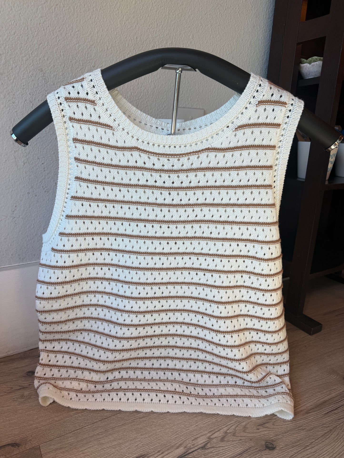 A sleeveless crochet knit top in cream and brown stripes, displayed on a black hanger against a neutral indoor wall and wood floor.