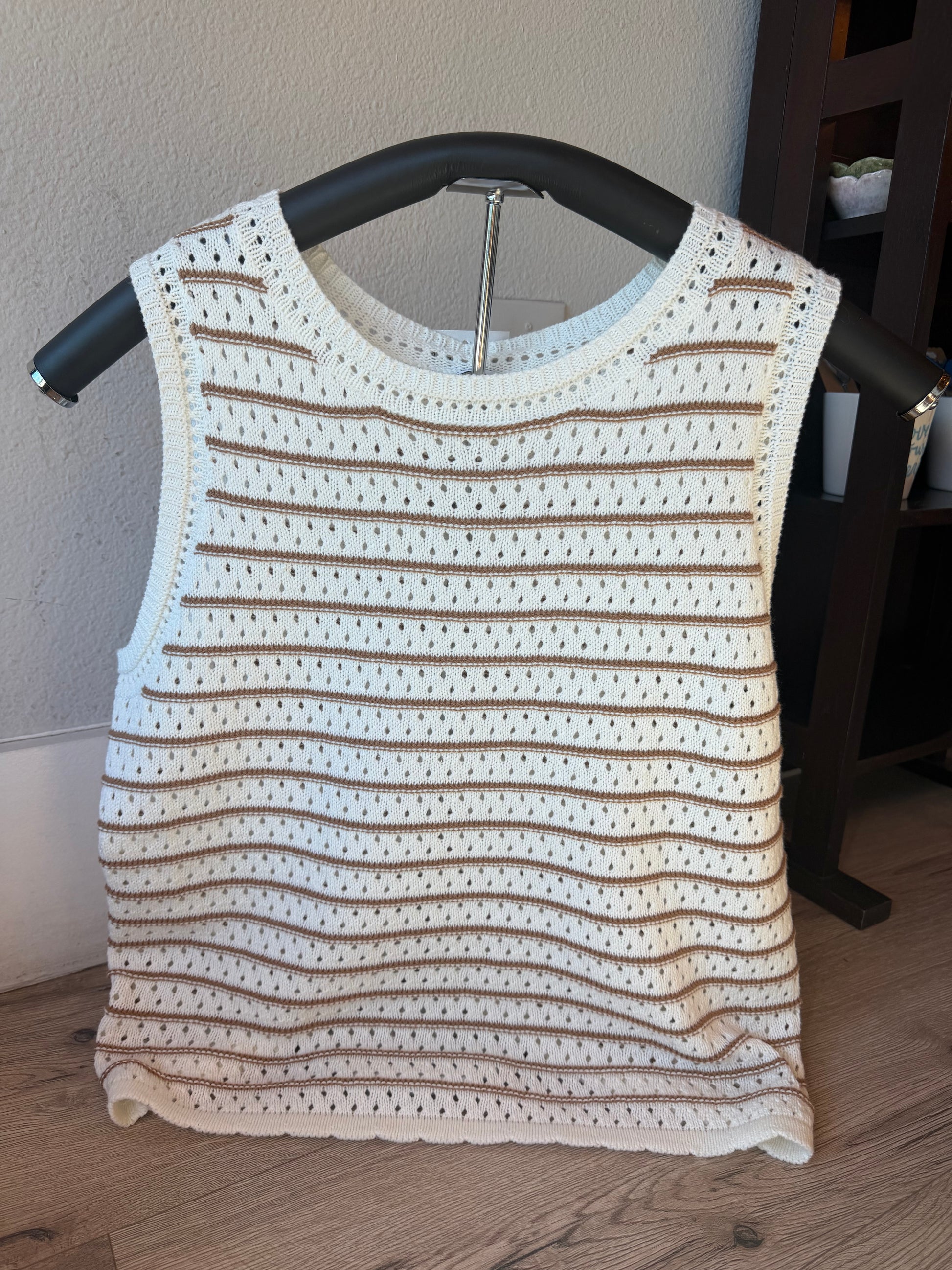 A sleeveless crochet knit top in cream and brown stripes, displayed on a black hanger against a neutral indoor wall and wood floor.