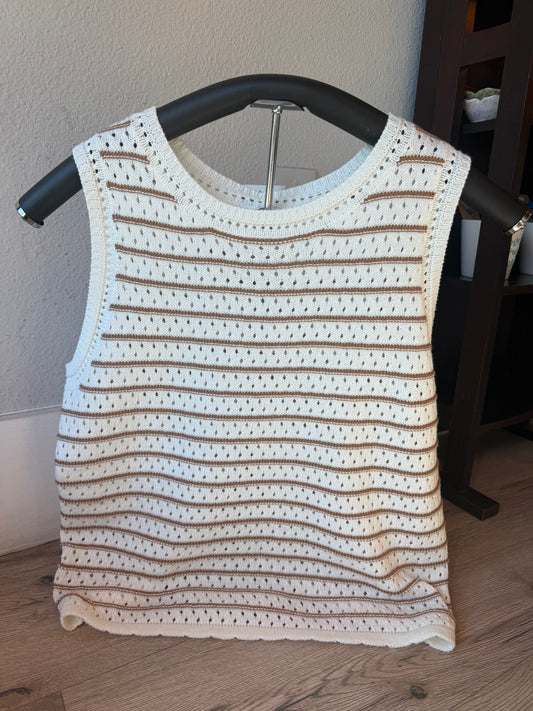 A sleeveless crochet knit top in cream and brown stripes, displayed on a black hanger against a neutral indoor wall and wood floor.