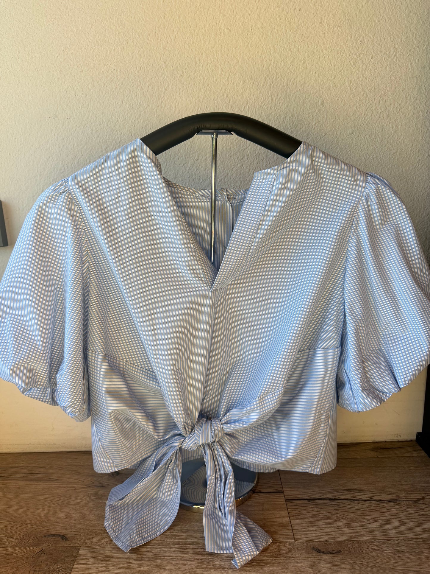 A light blue and white striped short-sleeve blouse with a V-neck and front tie knot, displayed on a black hanger against a beige wall and wood floor.