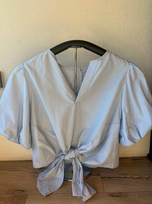 A light blue and white striped short-sleeve blouse with a V-neck and front tie knot, displayed on a black hanger against a beige wall and wood floor.