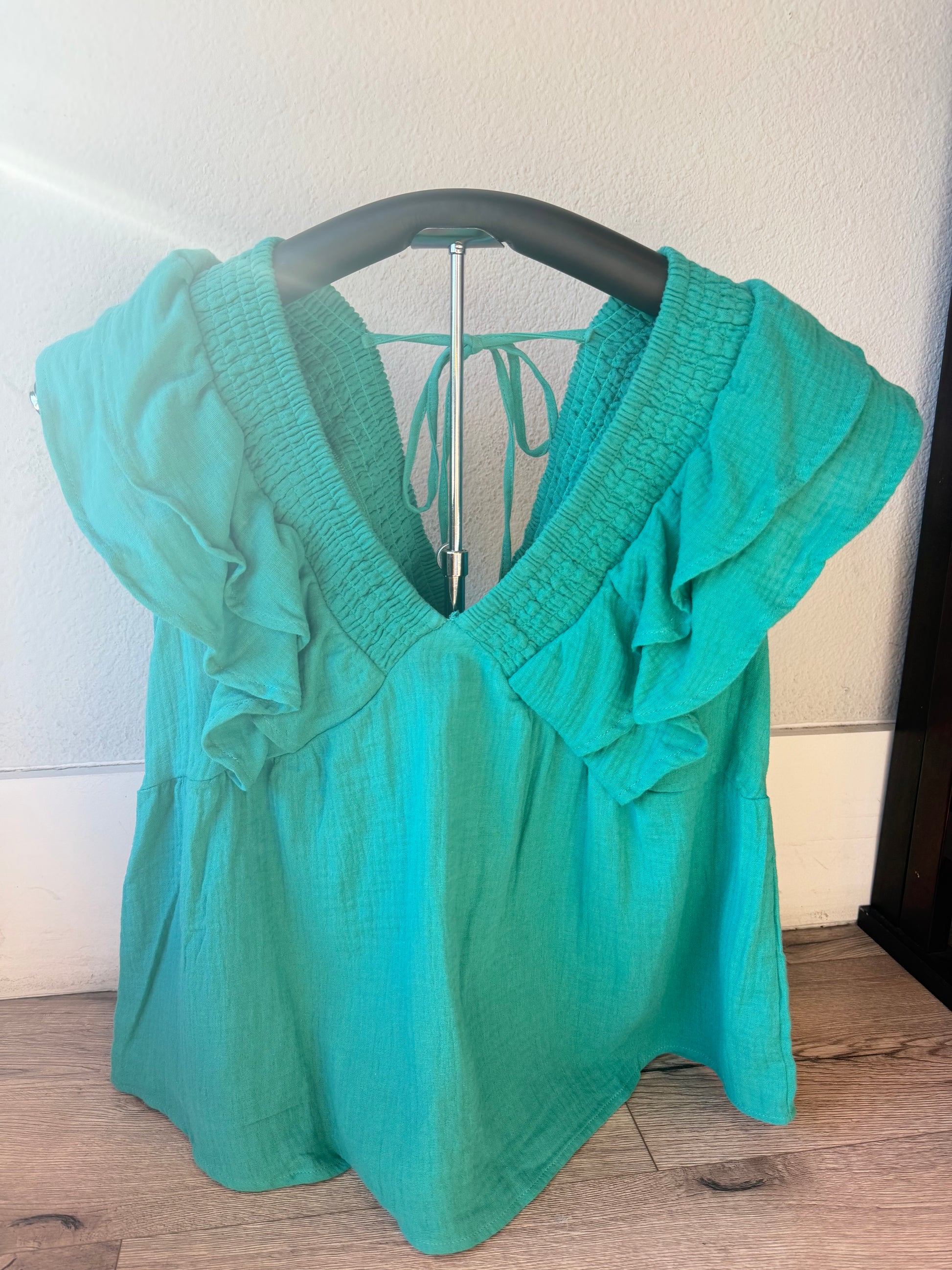A teal green short-sleeve blouse with ruffle details and a smocked V-neckline, hanging on a black hanger against a white wall and wood floor.