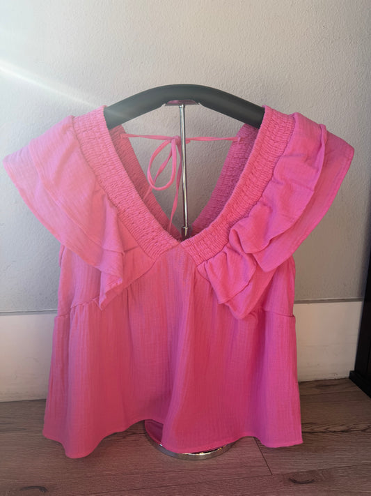 A bright pink short-sleeve blouse with ruffled trim and a tie detail at the back, displayed on a hanger against a plain indoor wall.