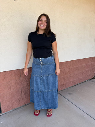 A woman stands outdoors against a beige and brown stucco wall, wearing a black short-sleeve t-shirt, a long tiered blue denim skirt, and red heels.