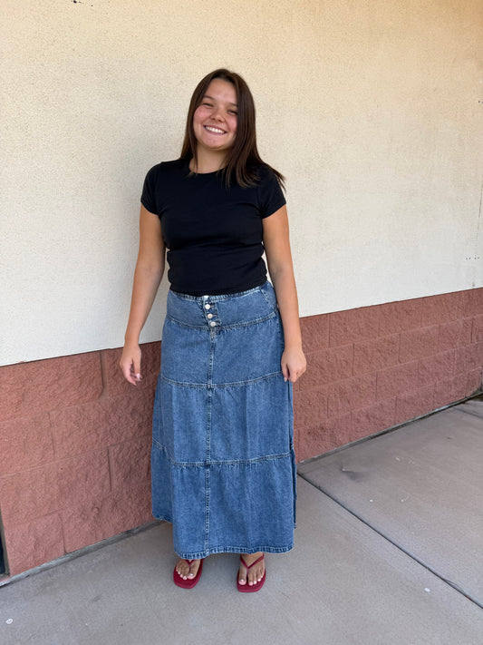 A woman stands outdoors against a beige and brown stucco wall, wearing a black short-sleeve t-shirt, a long tiered blue denim skirt, and red heels.