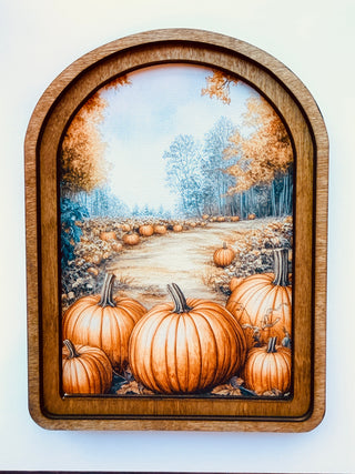 An arched wooden framed art print depicting a pumpkin patch scene with large orange pumpkins, a path, and trees in autumn colors, mounted on a plain white wall.