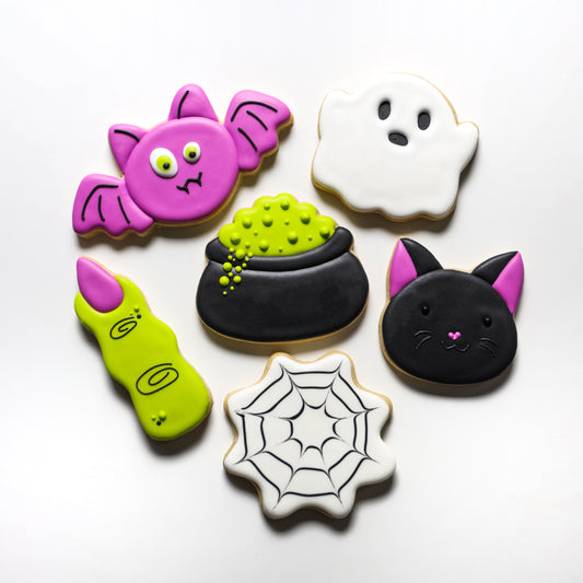 Halloween-themed cookies including a pink bat, white ghost, black cat, green potion, and spider web on a white background.