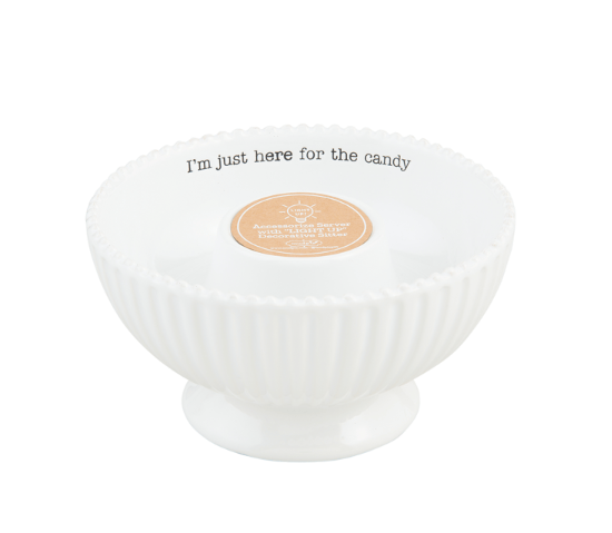 A white ceramic candy bowl with a fluted rim and pedestal base, featuring the printed phrase 'I'm just here for the candy' in black around the inner rim and a circular gold sticker in the center.