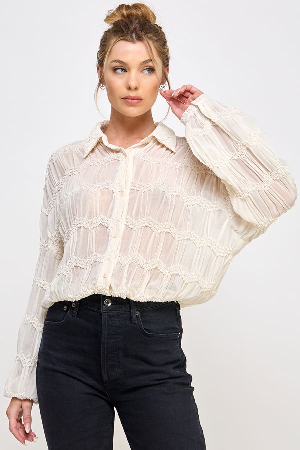 Woman wearing a sheer buttercream blouse with long sleeves and black pants on a light gray background