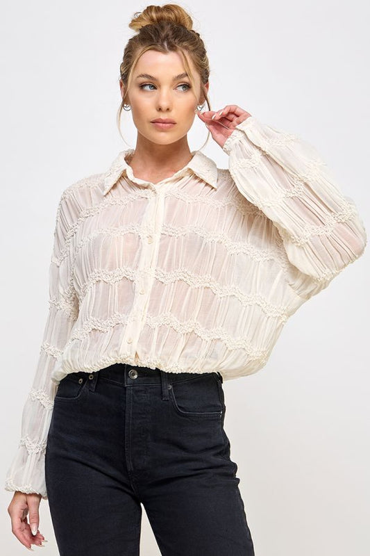Woman wearing a sheer buttercream blouse with long sleeves and black pants on a light gray background